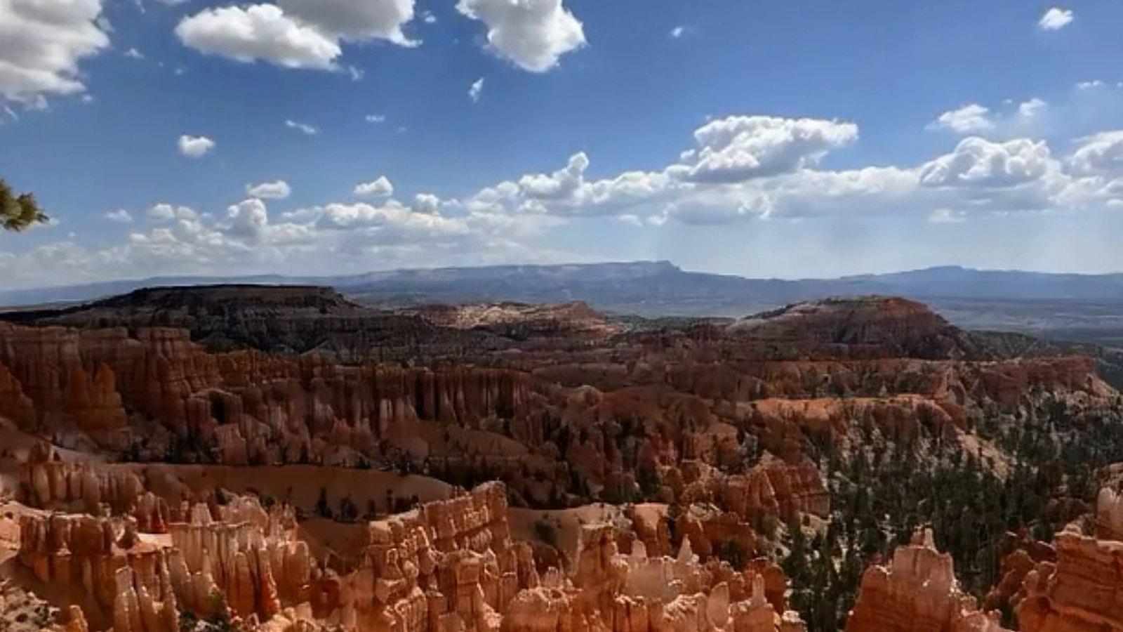 The Bryce Amphitheater seen from the rim: rows of orange and cream hoodoos and dark pines under a blue sky with scattered clouds, the plateau stretching to the horizon.