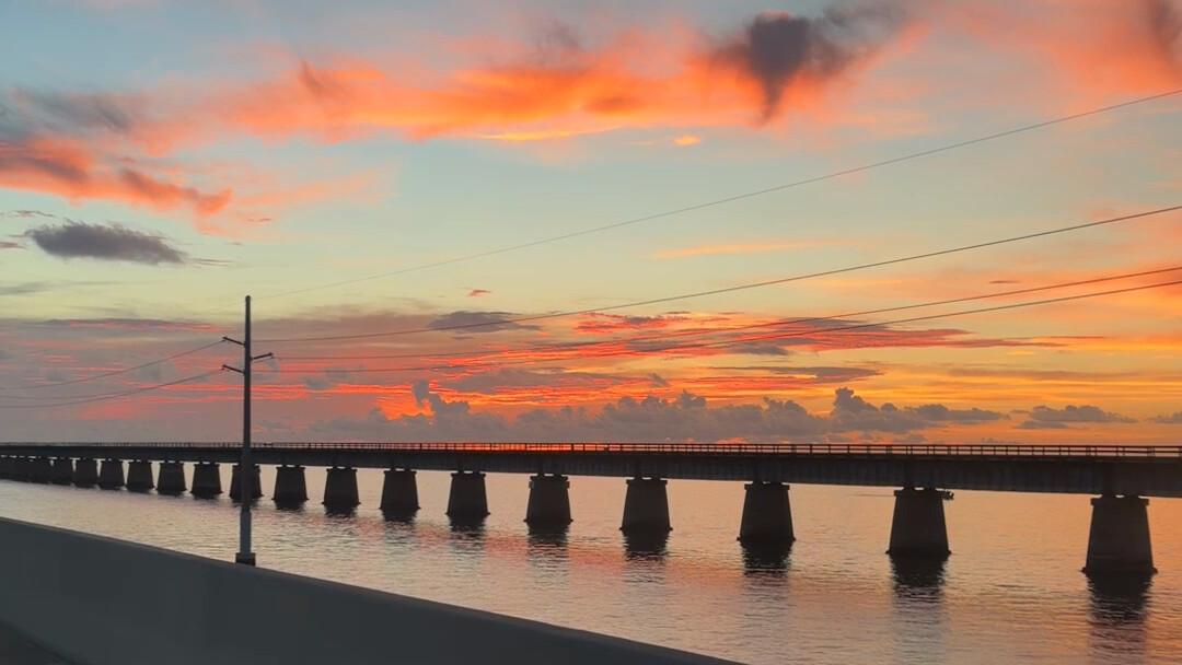 The old Seven Mile Bridge running across calm water under an orange and pink sunset sky, seen from the road on the new bridge.