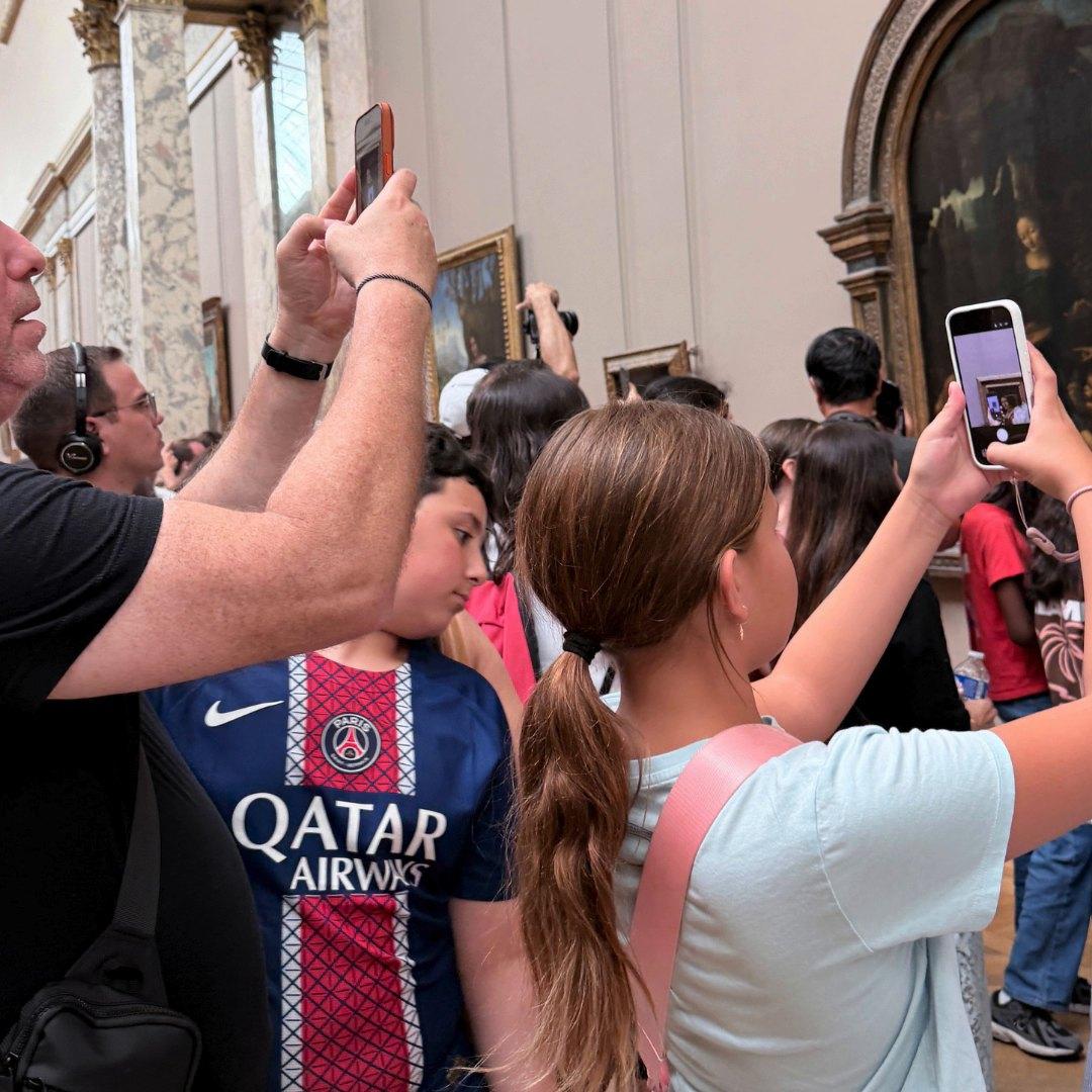 A tight crowd in a Louvre painting gallery holds phones up toward the pictures; two children in the foreground look up at the walls.