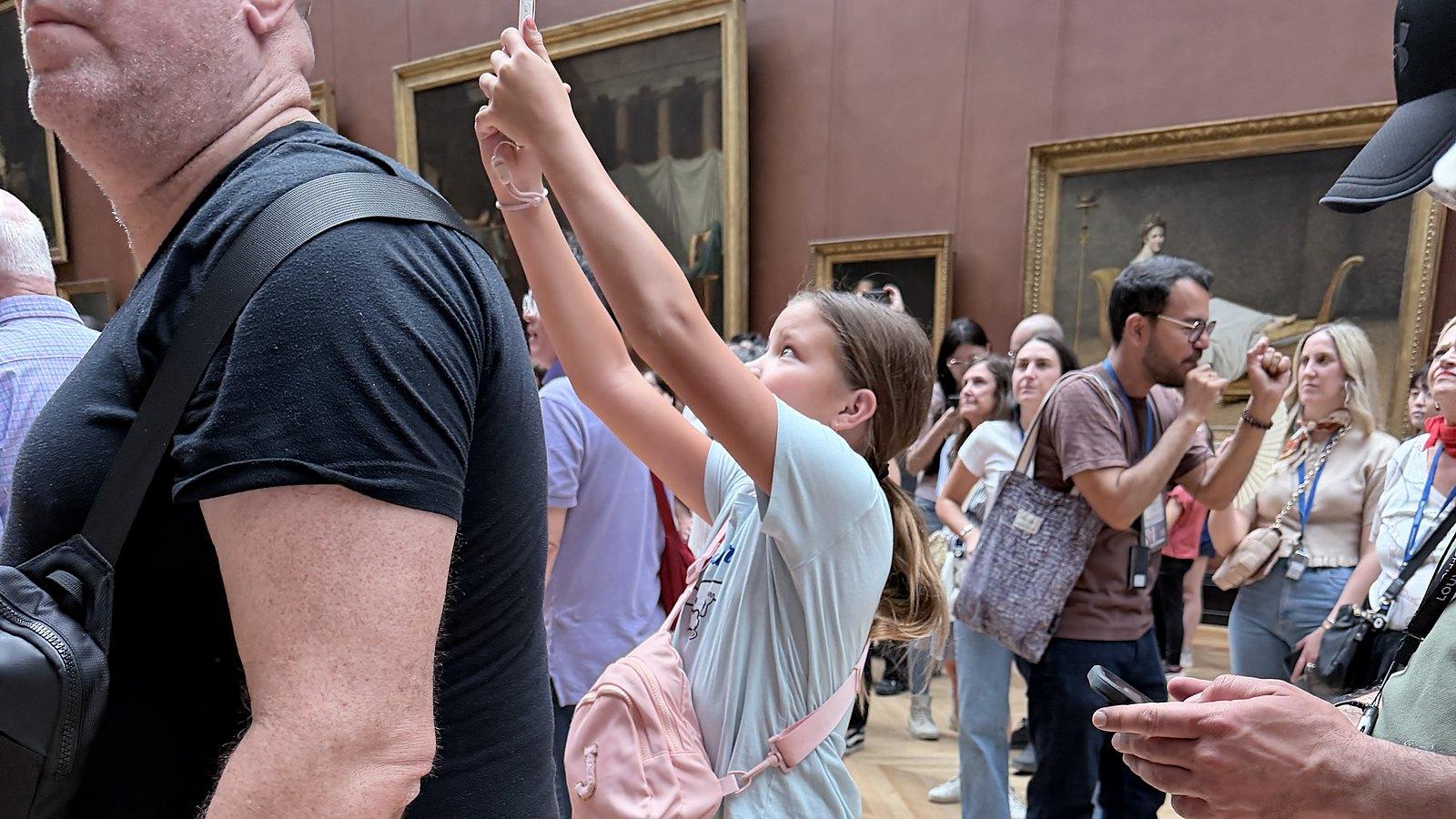 In a crowded, red-walled Louvre gallery, a girl with a pink bag raises her phone high to photograph over the heads of other visitors.