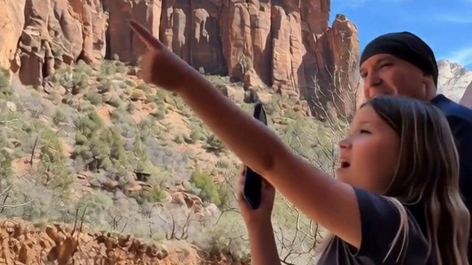 A girl on a canyon trail points up at towering red sandstone cliffs in Zion National Park, a parent beside her looking the same way.
